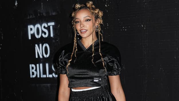 Singer Tinashe stands in front of a "Post No Bills" sign wearing a black cropped top with puff sleeves and matching pants, smiling for the camera