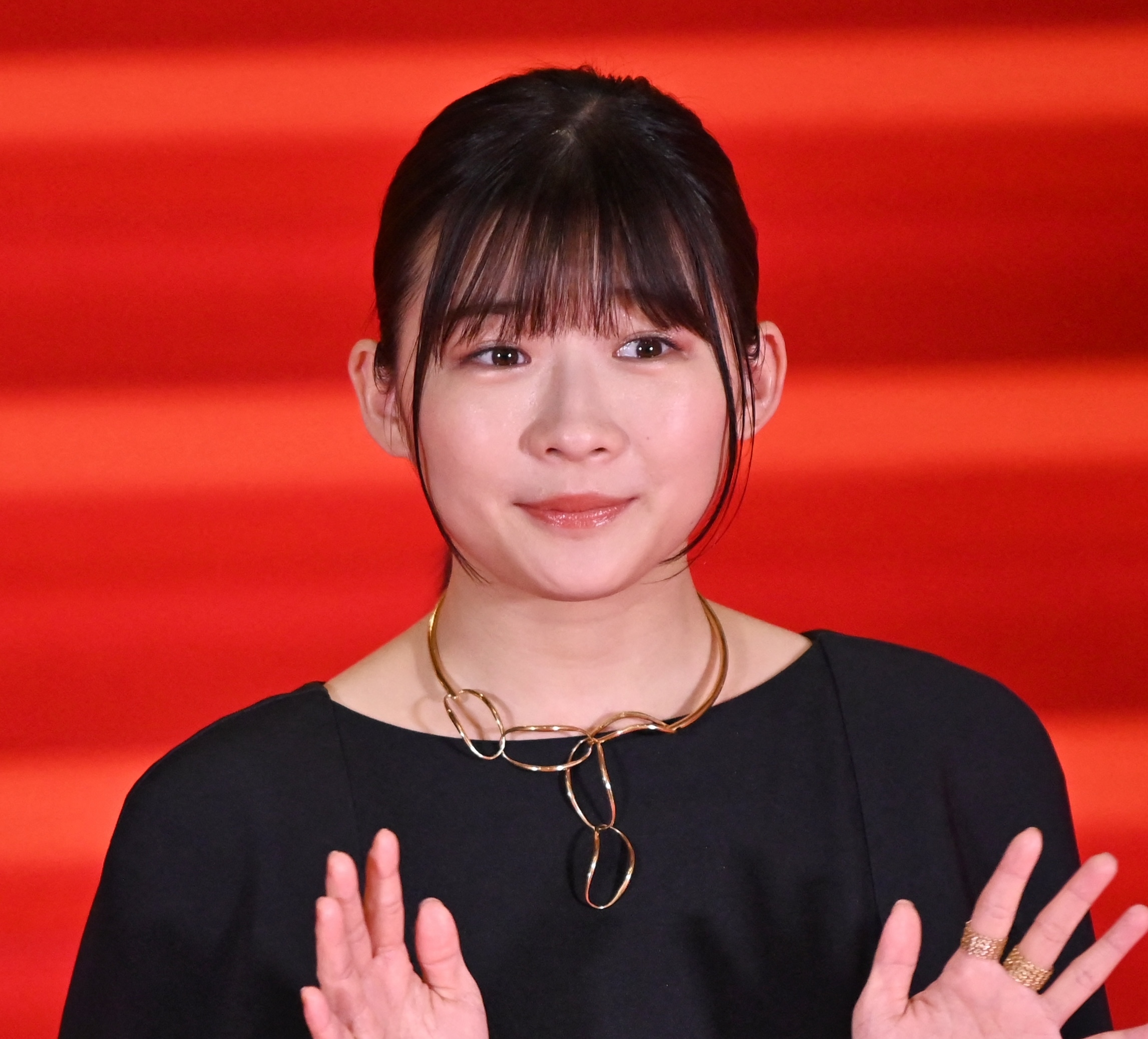 女性が黒いドレスと独特なネックレスを着用してレッドカーペットに立っています。