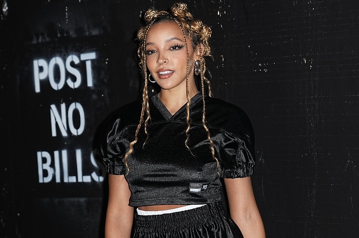 Tinashe standing in front of a wall with a "Post No Bills" sign, wearing a sporty black outfit with braided hair styled in Bantu knots