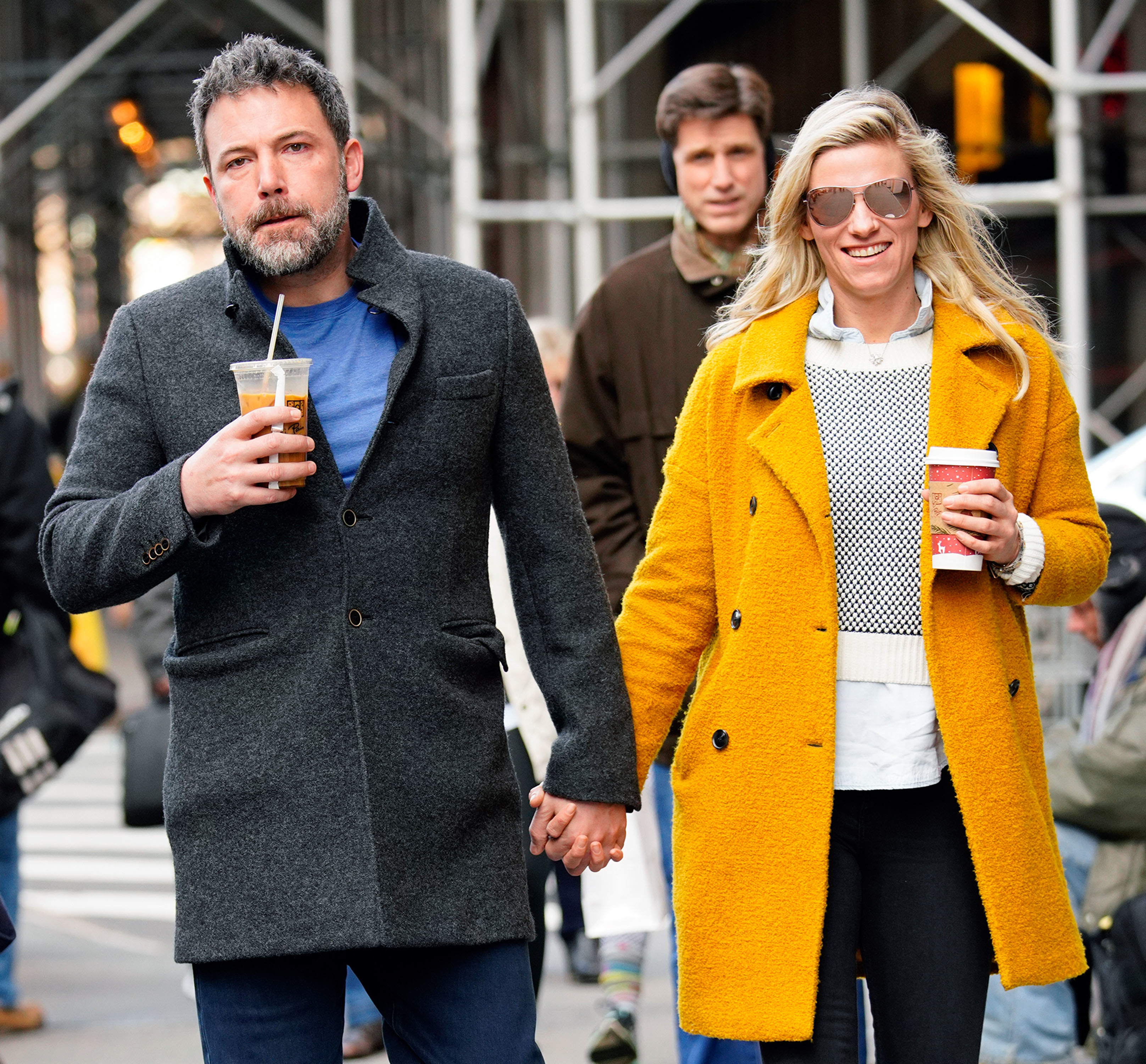 Ben Affleck in a dark coat holding iced coffee and Lindsay Shookus in a yellow coat and sunglasses holding hot coffee, walking hand in hand