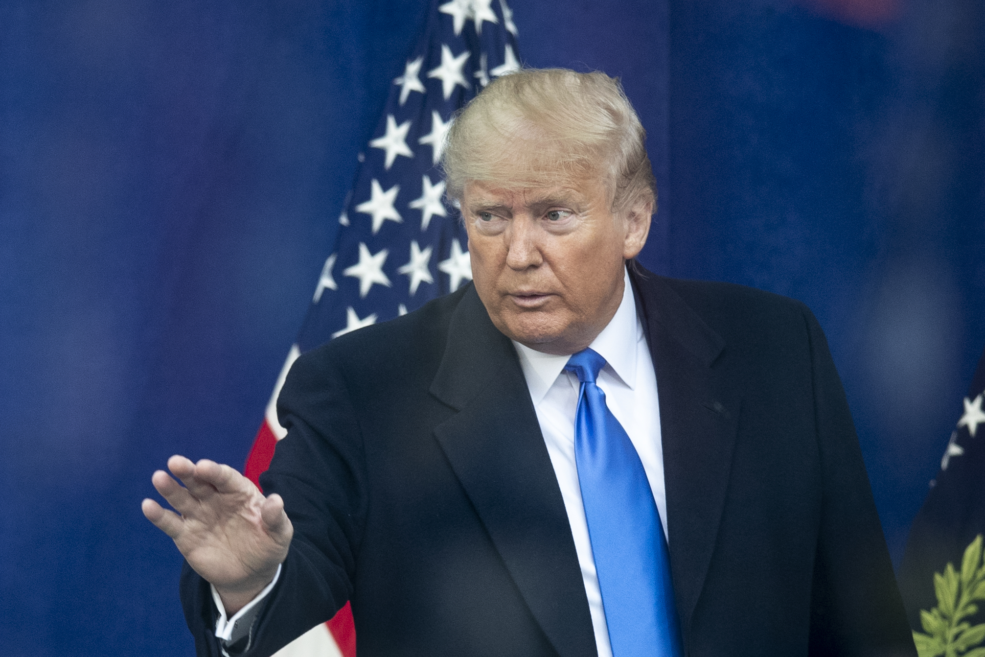 Donald Trump stands in front of an American flag, wearing a black overcoat and blue tie, with his right hand raised in a gesturing motion