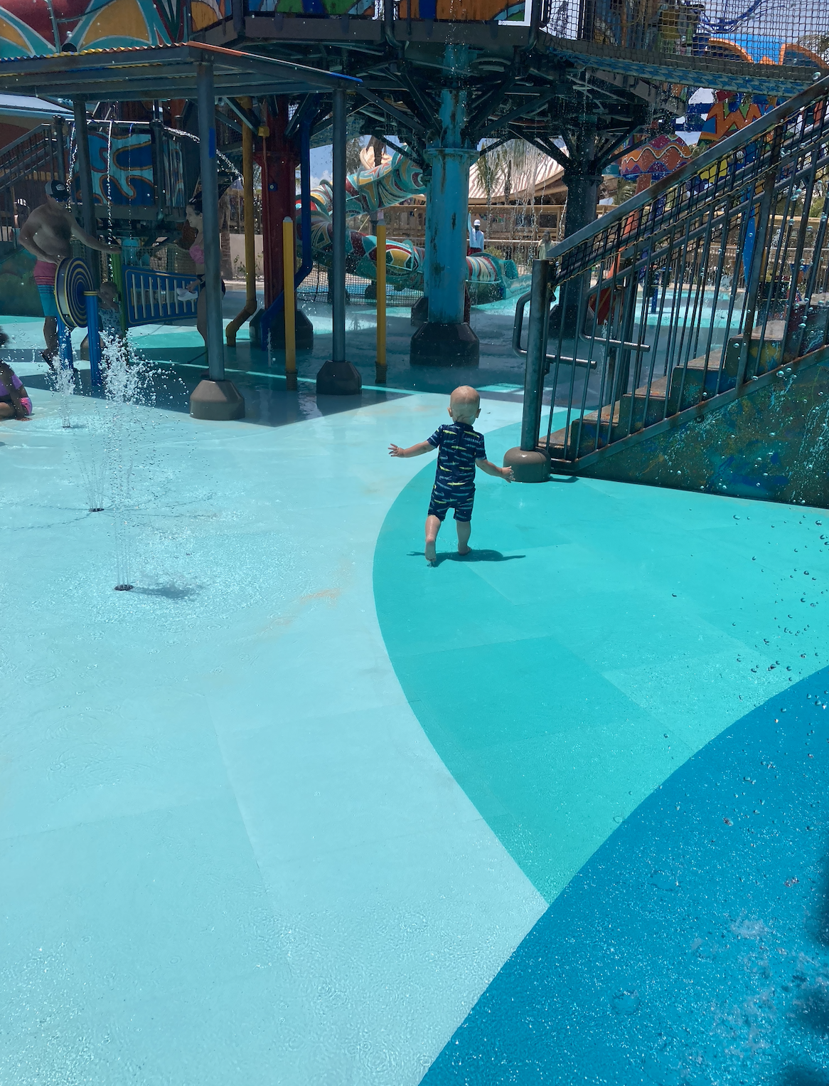 A toddler in a swimsuit is walking alone in a splash pad water play area with various water features and slides in the background