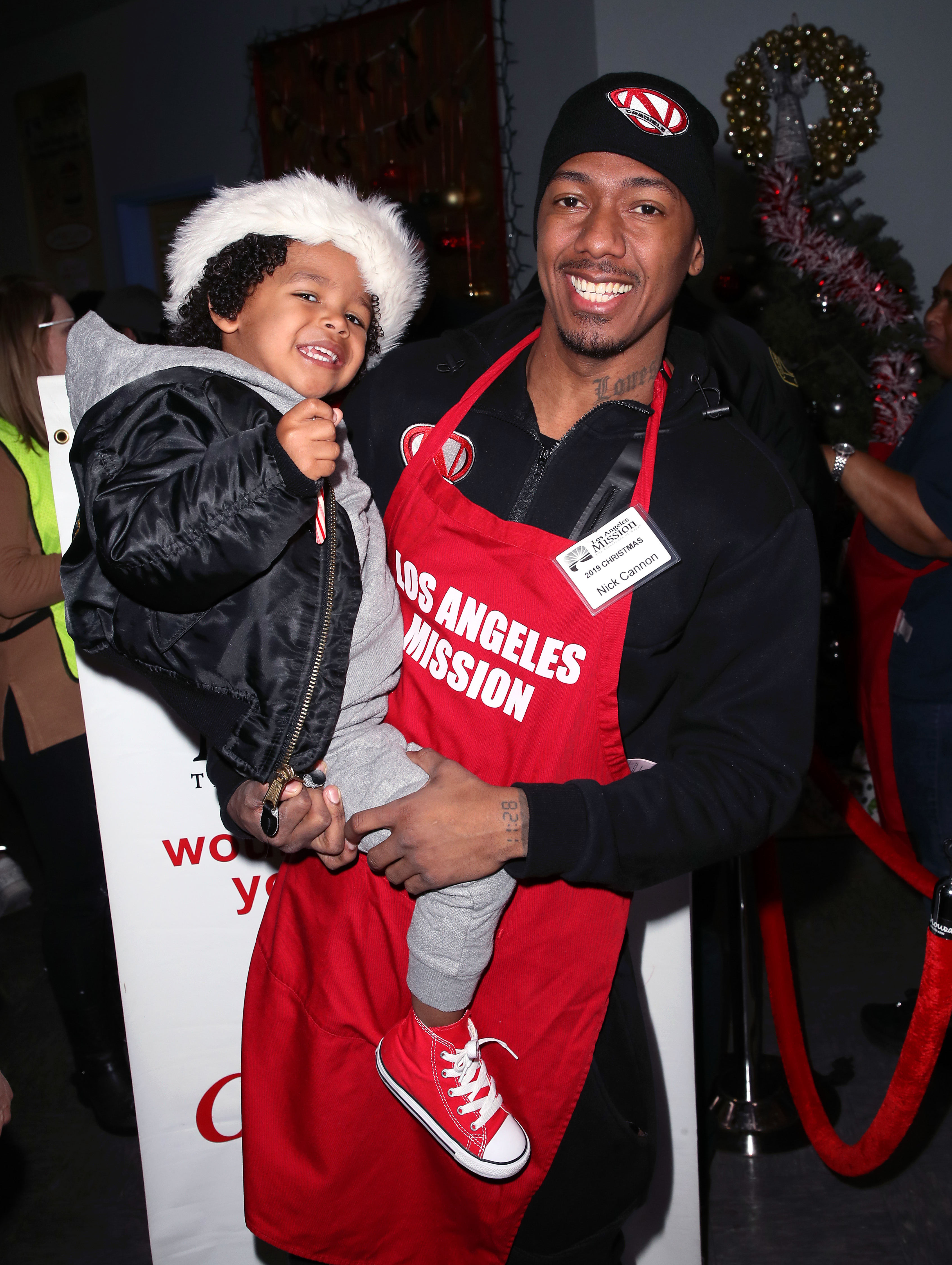 Nick Cannon holds a smiling child while wearing a red &quot;Los Angeles Mission&quot; apron at a charity event