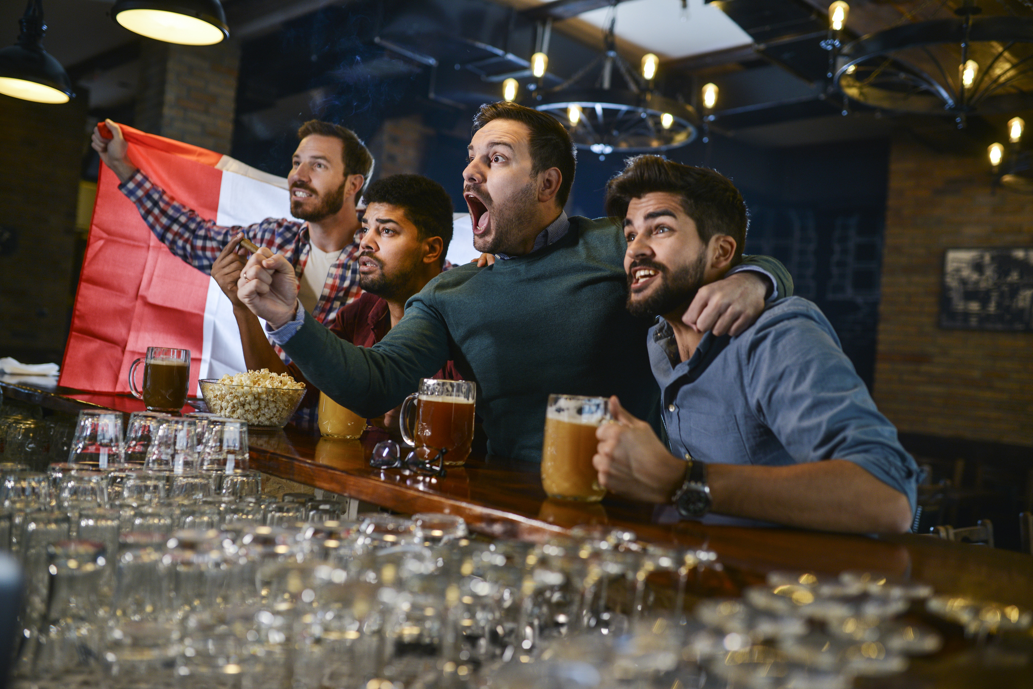 A group of four friends cheers and celebrates at a bar while watching a sports game, one holding a flag. Drinks and snacks are on the bar counter in front of them