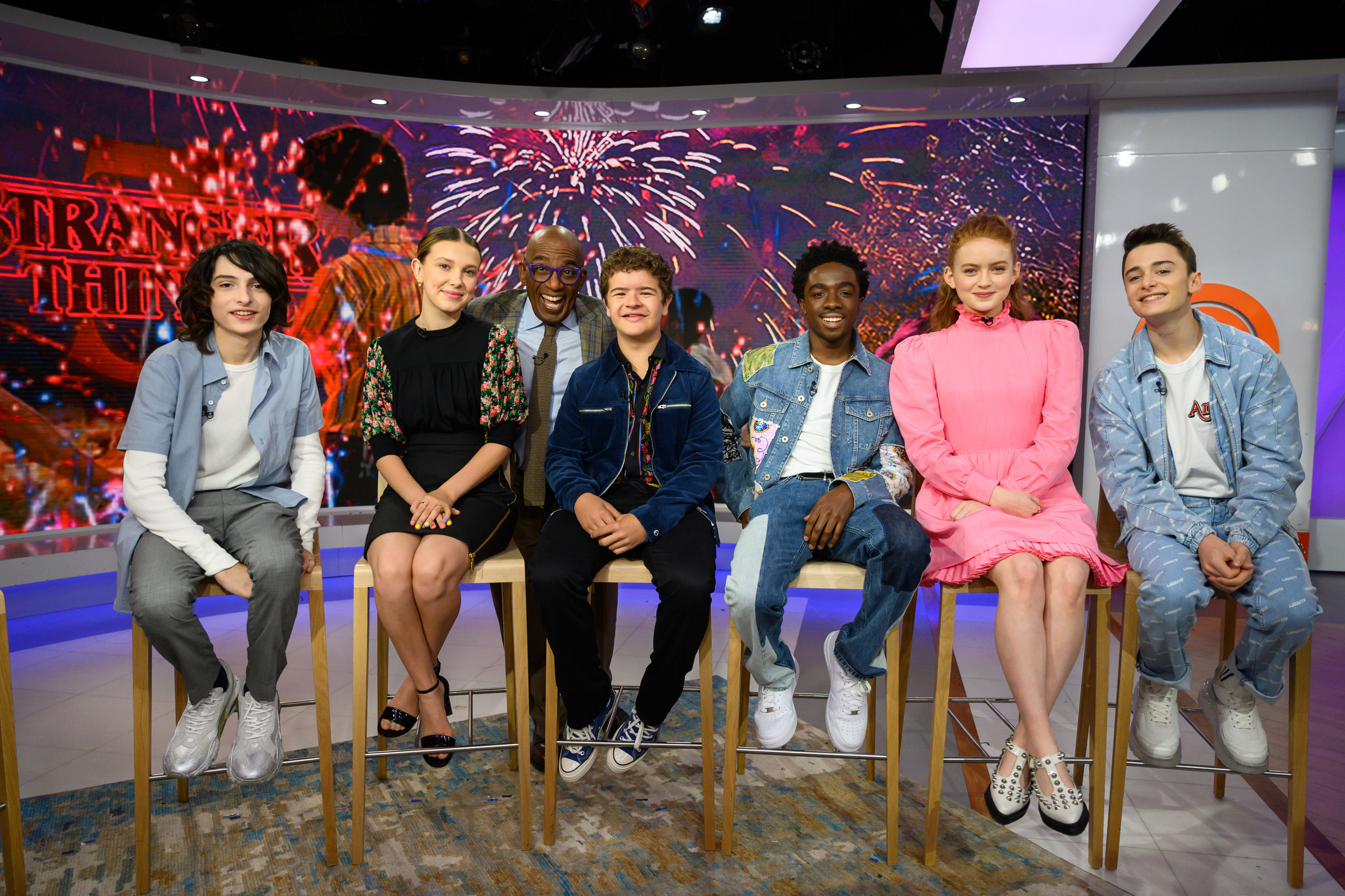 Finn Wolfhard, Millie Bobby Brown, Al Roker, Gaten Matarazzo, Caleb McLaughlin, Sadie Sink, and Noah Schnapp sit on stools on a TV show set with fireworks in the background