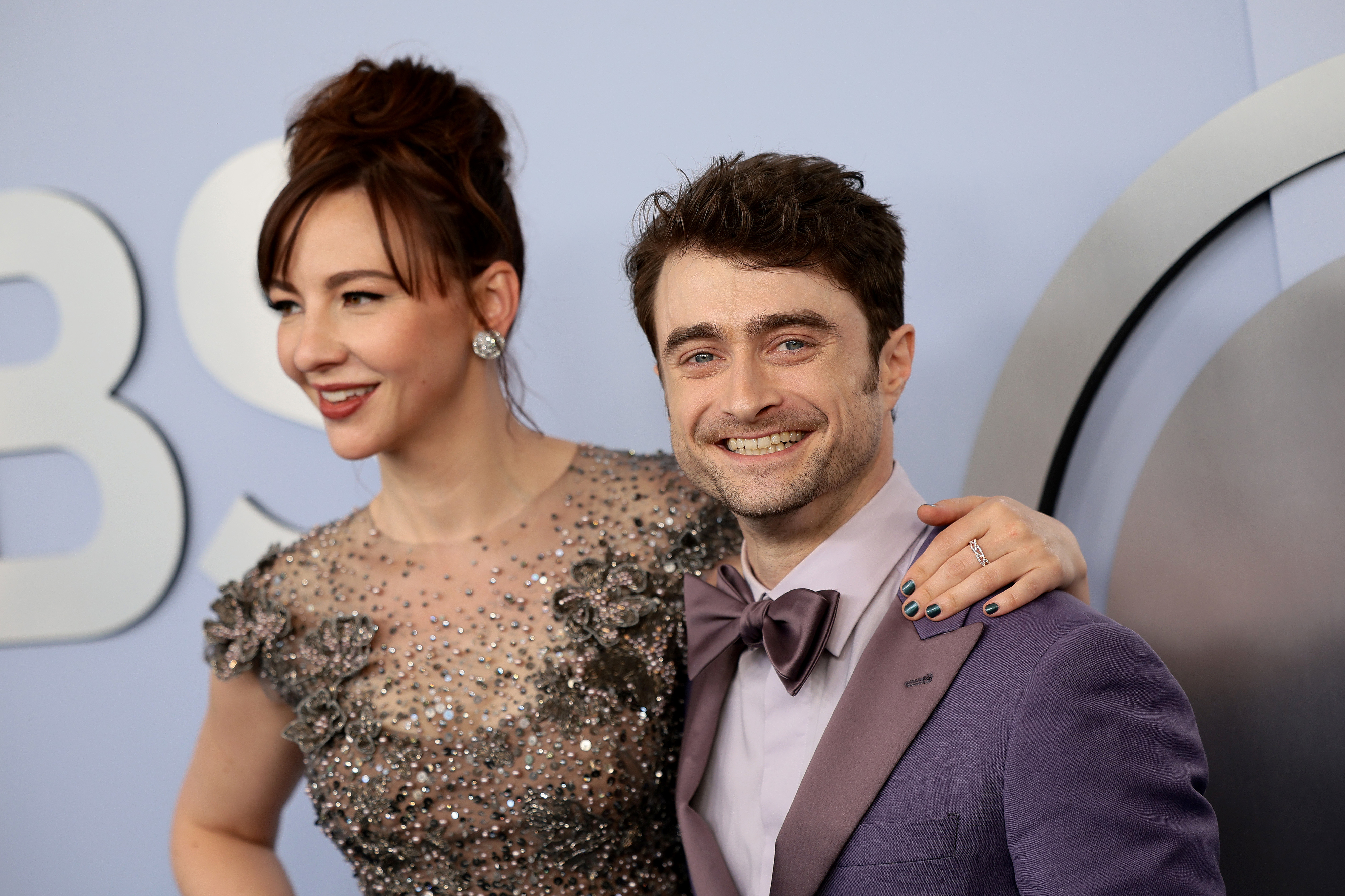 Daniel Radcliffe is smiling and wearing a suit with a bow tie, while Erin Darke next to him is wearing an embellished dress. Both are posing for a photo on the red carpet
