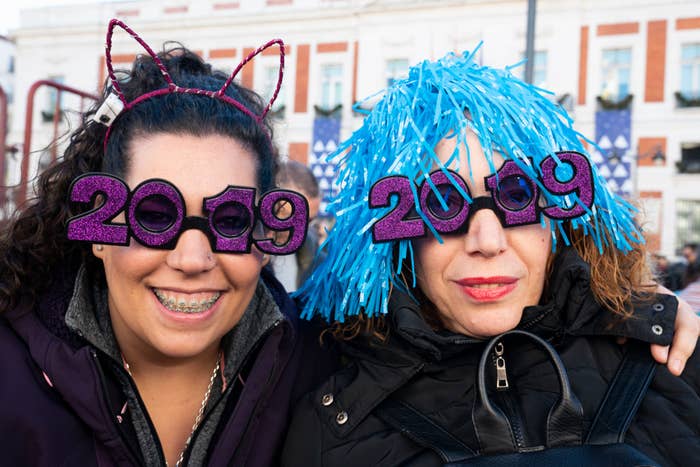 Two women wearing &quot;2019&quot; glasses, one with cat ear headband and the other with a blue wig, celebrate New Year's Eve outdoor
