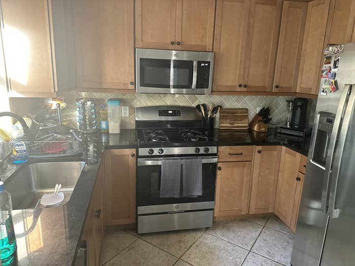 Modern kitchen with wooden cabinets, stainless steel appliances, a gas stove, a microwave, and a refrigerator. Counters have various kitchen items and a dish drying rack