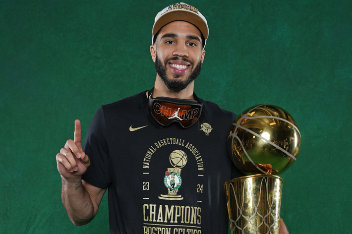 Jayson Tatum holding an NBA Championship trophy and wearing a "Champions" t-shirt, cap, and championship goggles, smiling and pointing upward with one finger