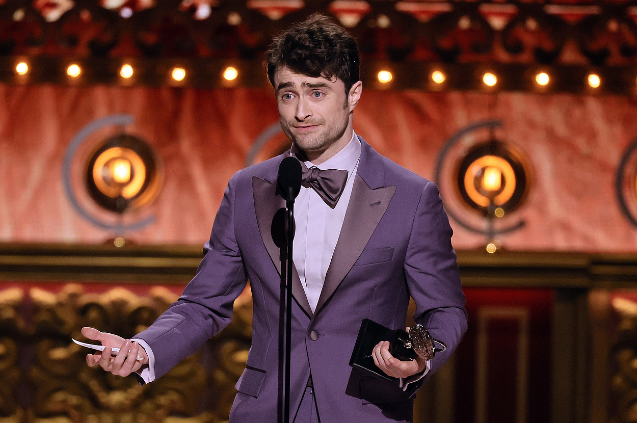 Daniel Radcliffe stands on stage in a suit, holding a microphone and an award, addressing the audience