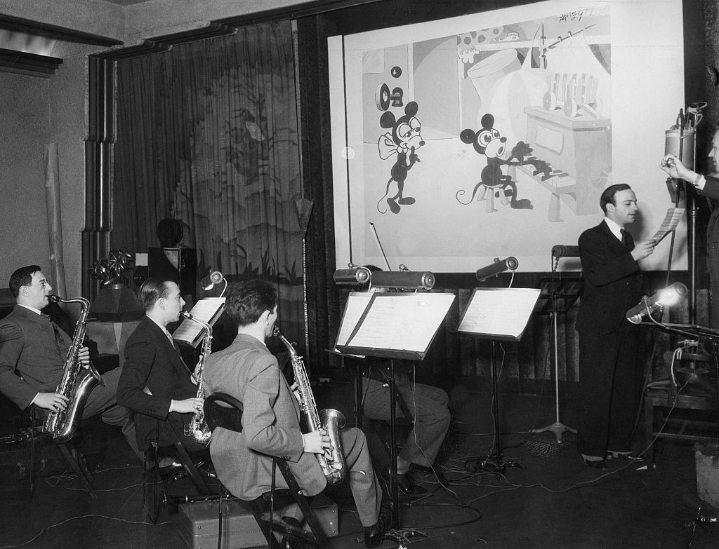 Several musicians play saxophones in a studio while Walt Disney oversees an animation of Mickey Mouse and Minnie Mouse projected on the screen behind them