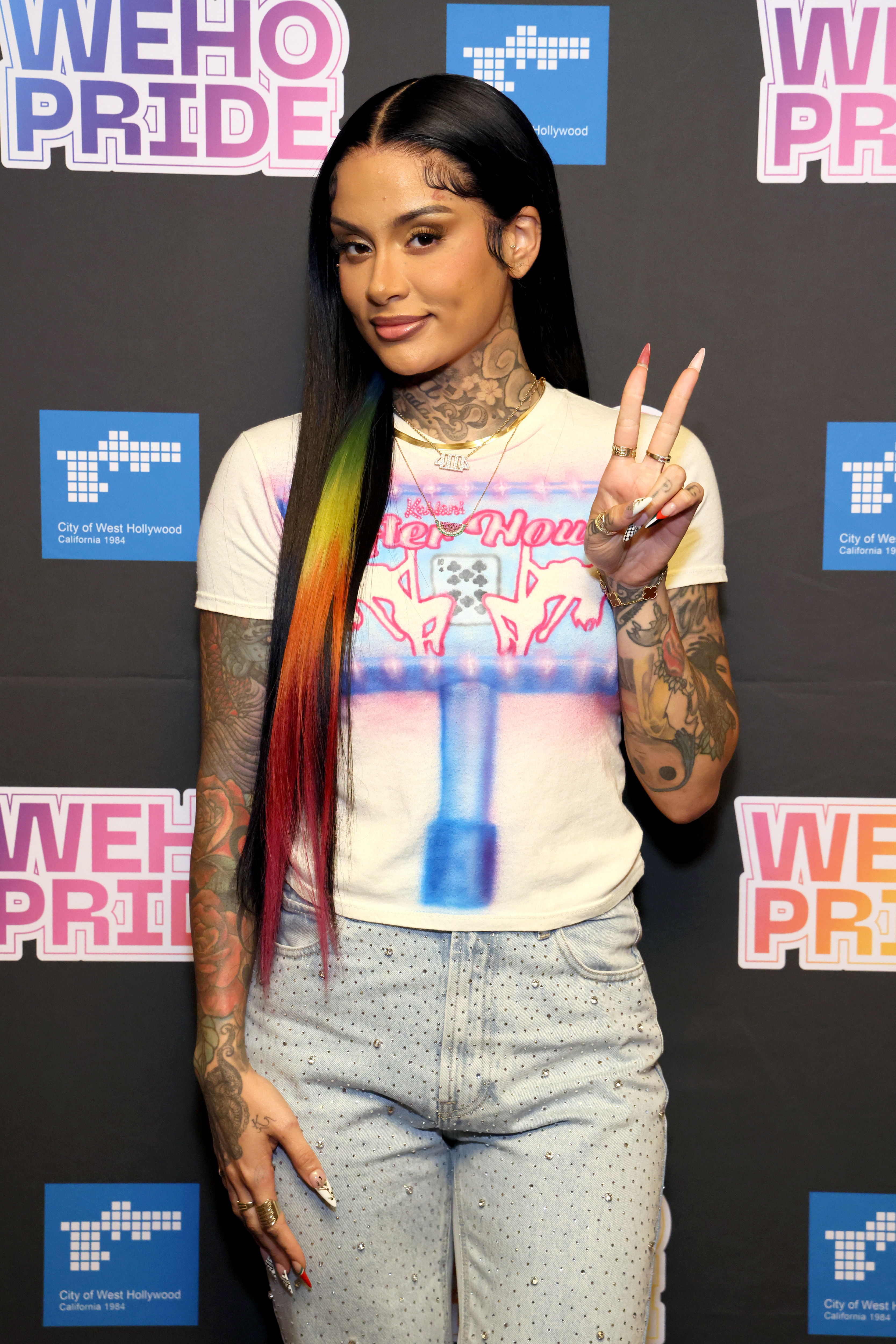 Kehlani at an event wearing a graphic tee, high-waisted jeans, and colorful hair, posing with a peace sign