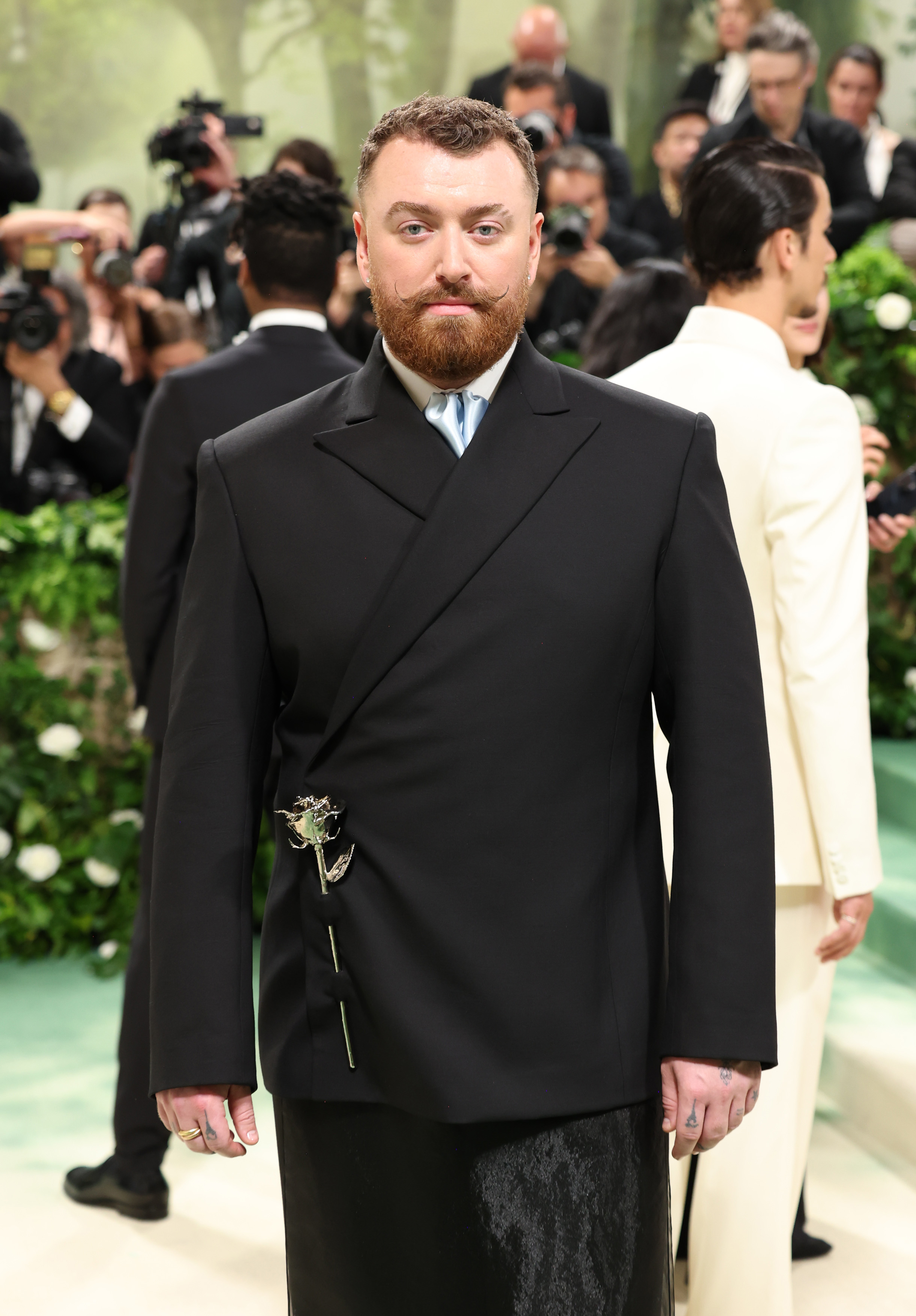 Sam Smith wearing a tailored black suit with a floral brooch on a formal red carpet event, surrounded by photographers