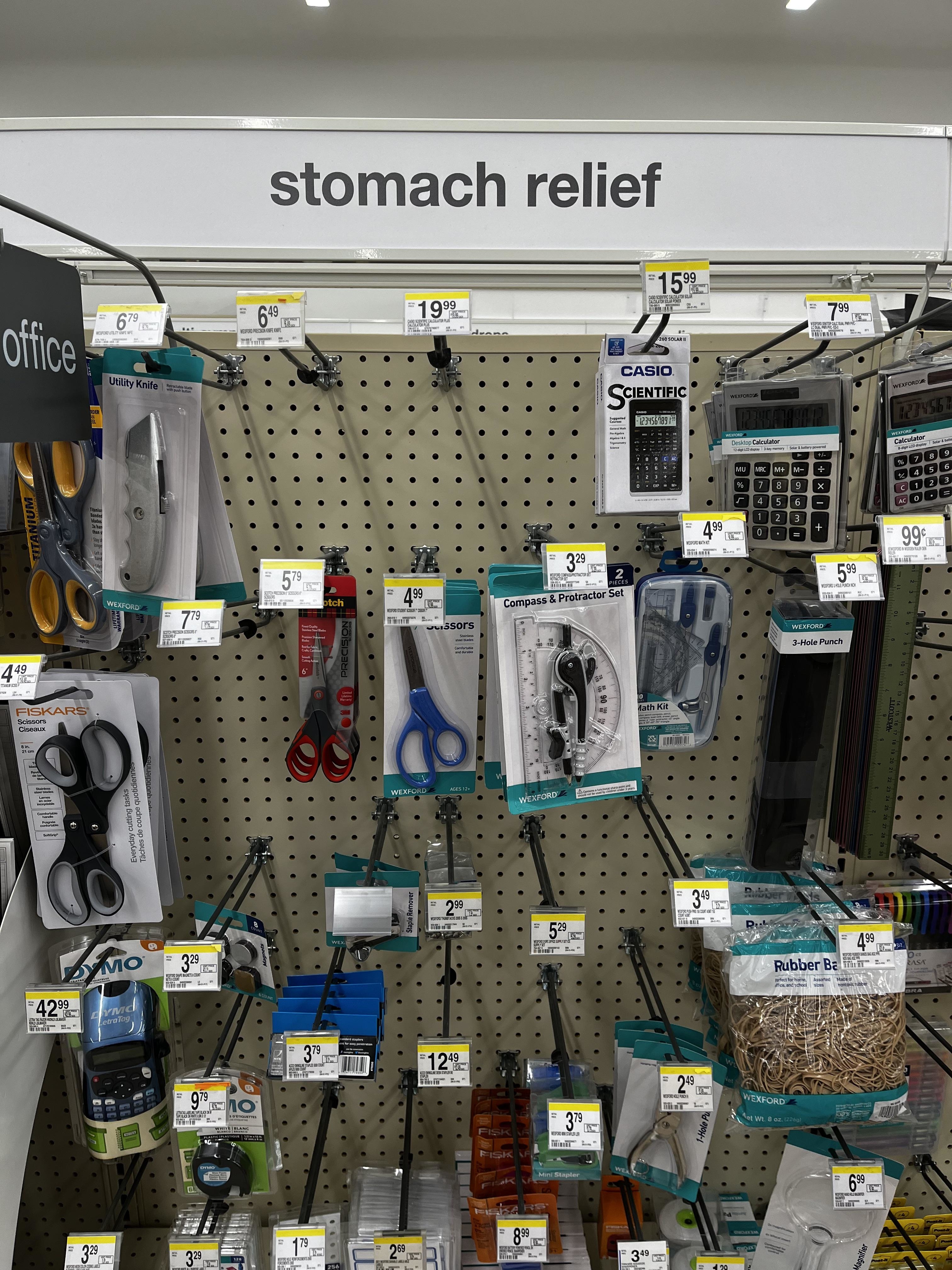 A store shelf labeled "stomach relief" mistakenly stocked with office supplies, including scissors, calculators, and staplers