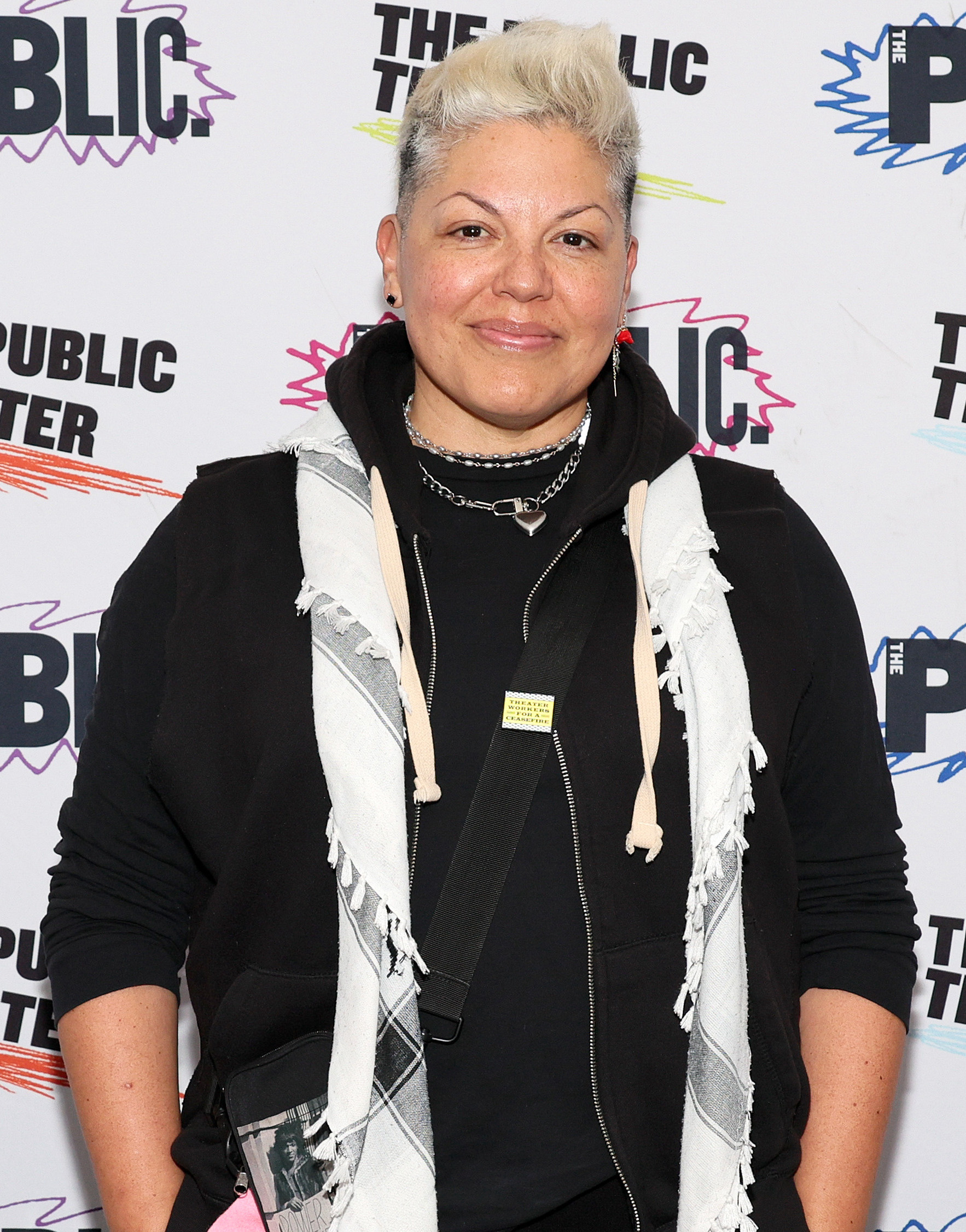 Sara Ramirez at The Public Theater event, wearing a checkered scarf, black hoodie, and crossbody bag