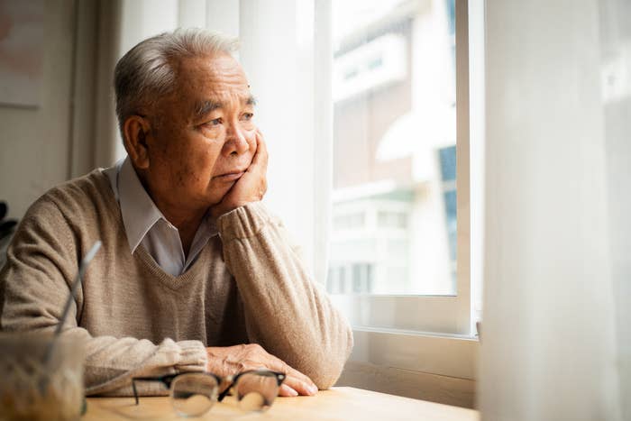 Elderly man sits by a window, resting his head on his hand, looking thoughtful and contemplative. A pair of glasses and a beverage sit on the table before him