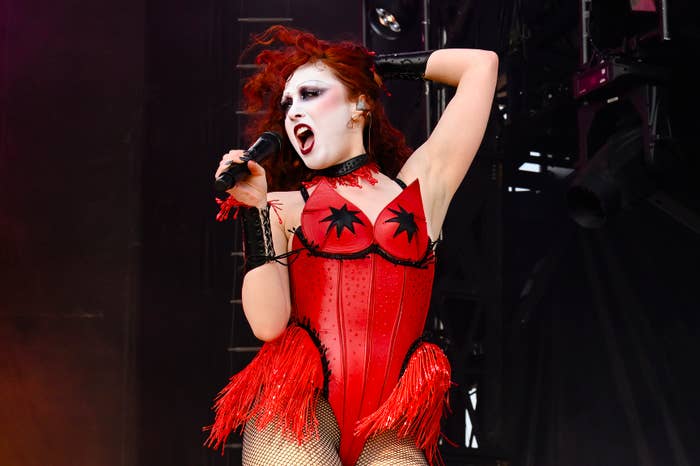 A performer with dramatic makeup and red fringe corset sings passionately while holding a microphone on stage
