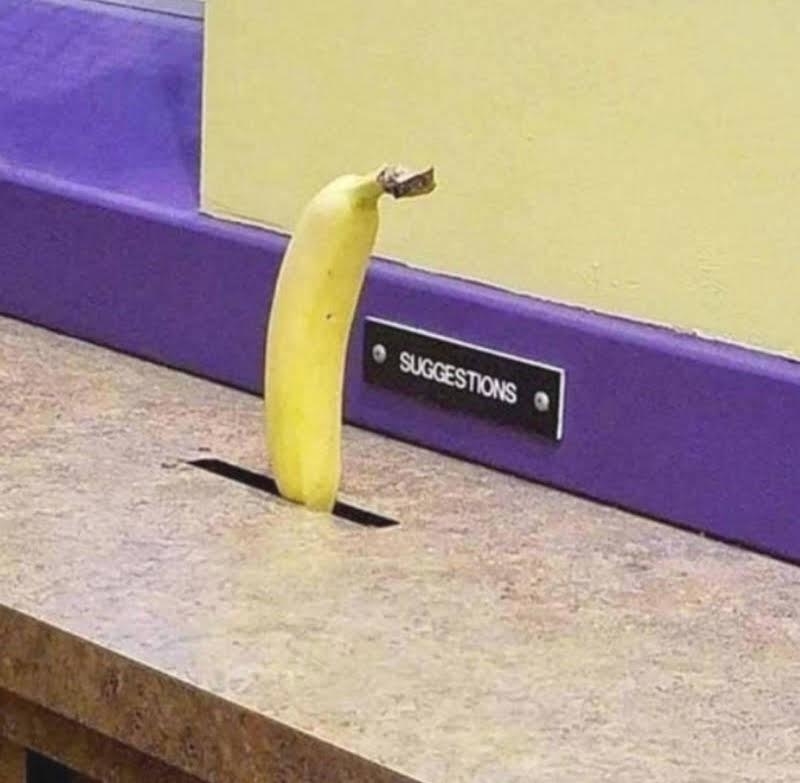 A banana is humorously placed in a suggestion box on a counter. The suggestion box is labeled "Suggestions."