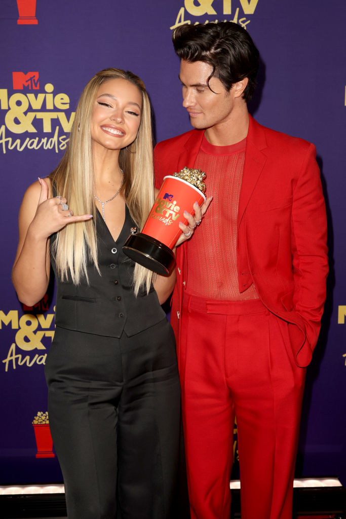 Madelyn Cline in a vest and pants and Chase Stokes in a suit with sheer shirt, posing with an award at the MTV Movie &amp;amp; TV Awards. Madelyn holds the award and smiles