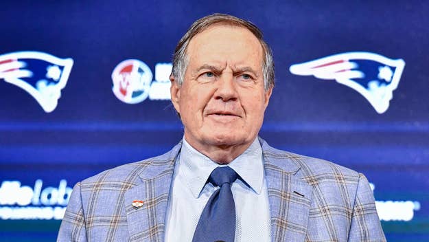 Bill Belichick stands in front of a backdrop bearing the New England Patriots logo, wearing a plaid suit jacket and tie