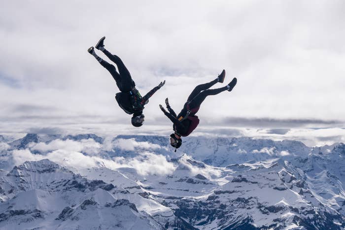 Two skydivers are performing acrobatic stunts mid-air with a backdrop of snow-capped mountain peaks below