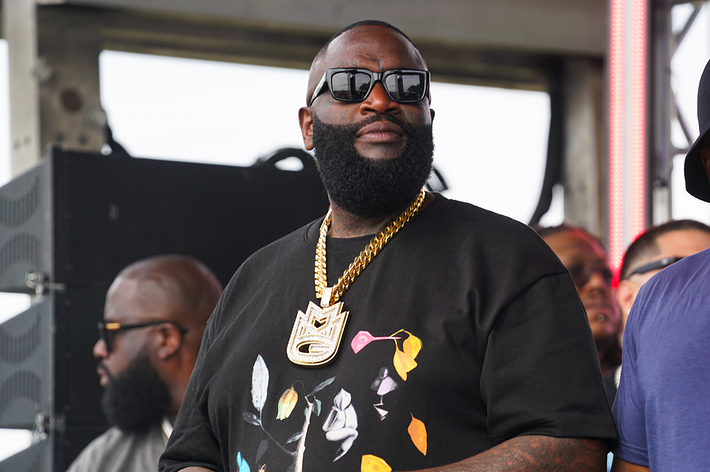 Rick Ross wearing a black graphic t-shirt and gold chain, standing on stage with a group of people at a music event