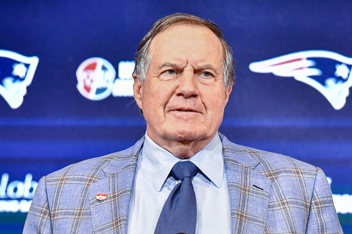 Bill Belichick stands in front of a Patriots backdrop, wearing a checkered blazer, button-up shirt, and tie