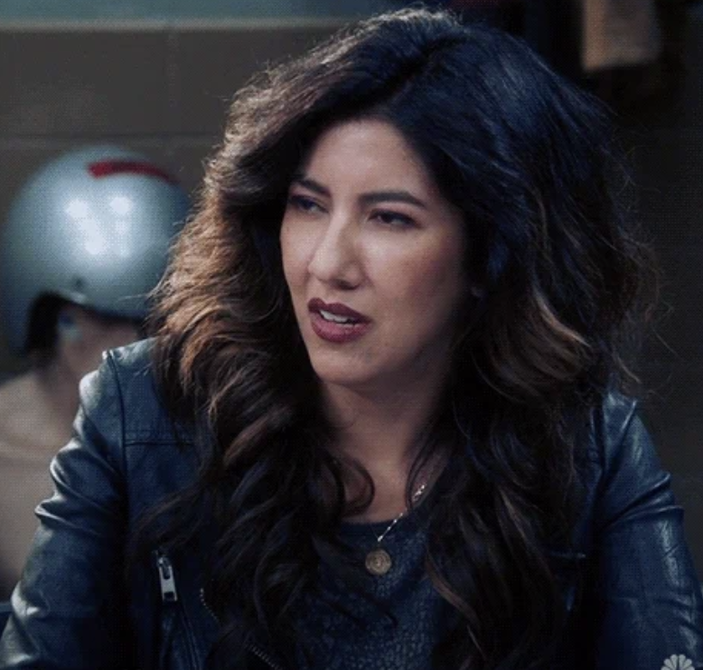 Stephanie Beatriz, dressed in a black leather jacket, appears in a scene from the TV show Brooklyn 99. She has long wavy hair and wears a pendant necklace