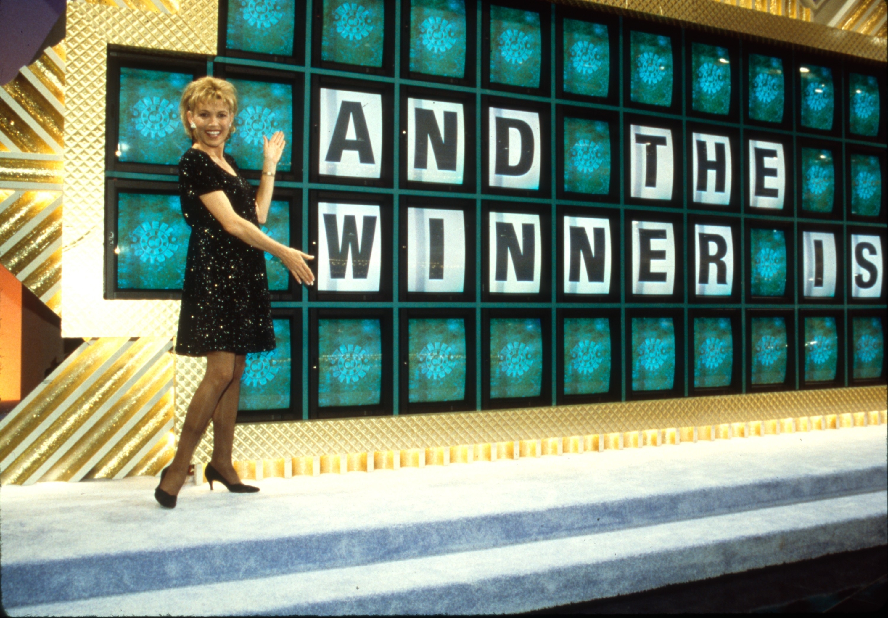 Vanna White in a sparkling dress gestures towards a game board on "Wheel of Fortune" that reads "AND THE WINNER IS"