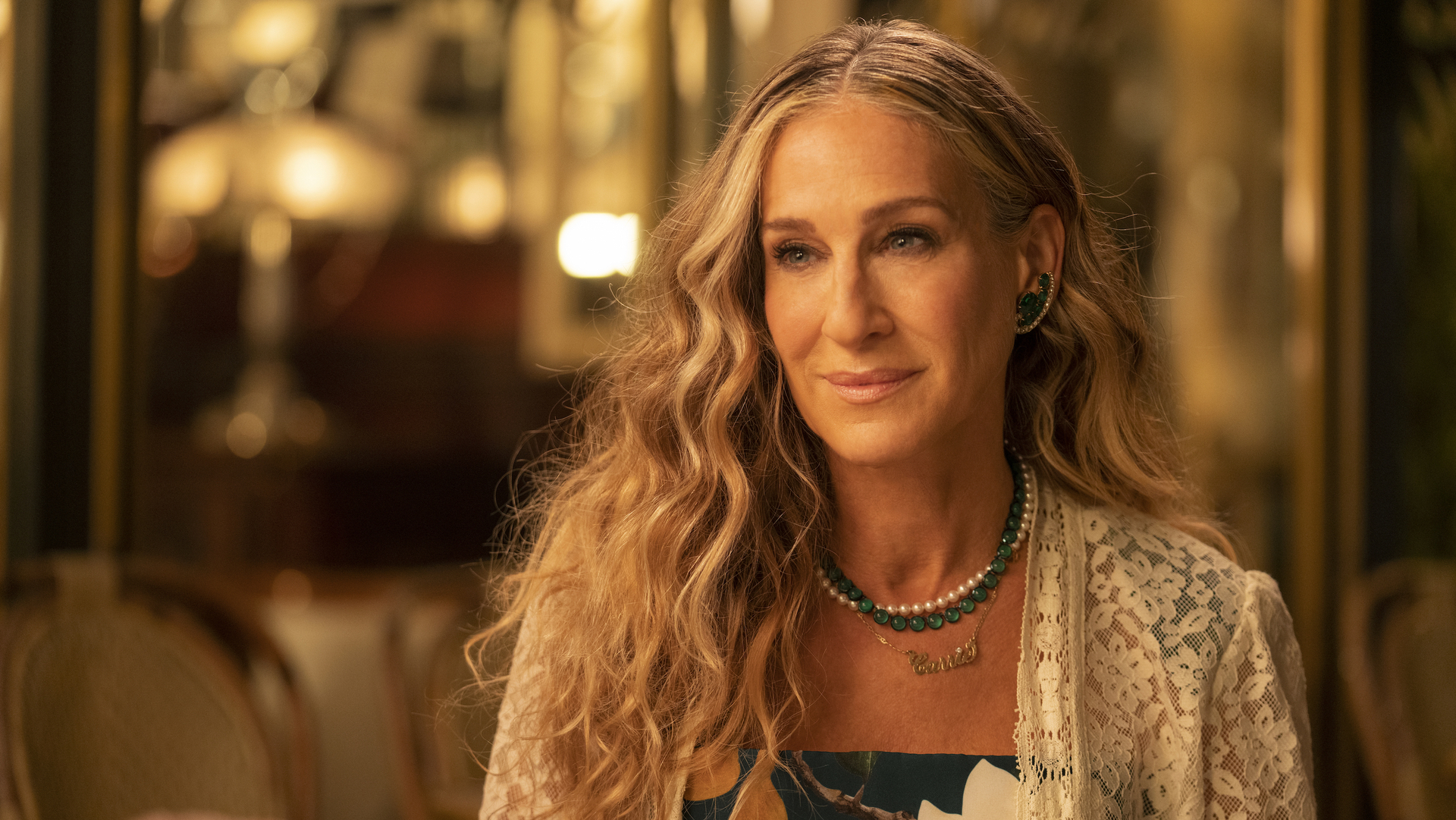 Sarah Jessica Parker wears a lace shawl over a patterned dress, with curly hair, pearl necklace, and earrings, sitting in a warmly lit setting