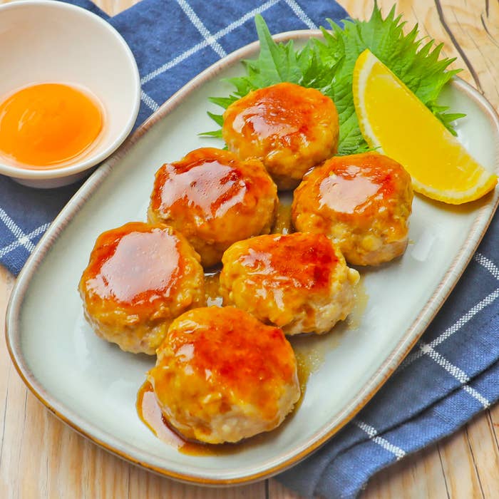 大皿に盛られた鶏つくねの照り焼きソースがかかった料理。横にレモンのくし切りと薬味が添えられている。