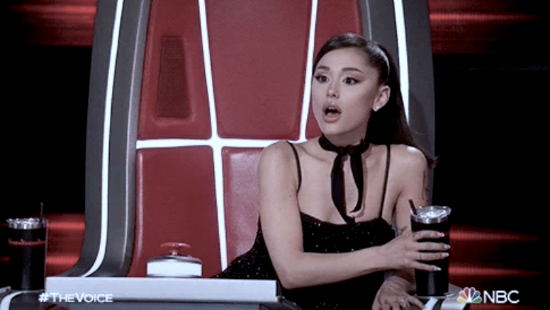 Ariana Grande, in a black strapless dress with a bow at her neck, sits on &quot;The Voice&quot; judge panel holding a drink and appears surprised