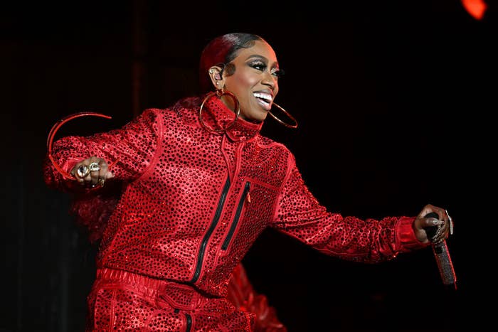 Missy Elliott performs on stage, smiling and wearing a stylish red studded tracksuit and large hoop earrings, holding a microphone