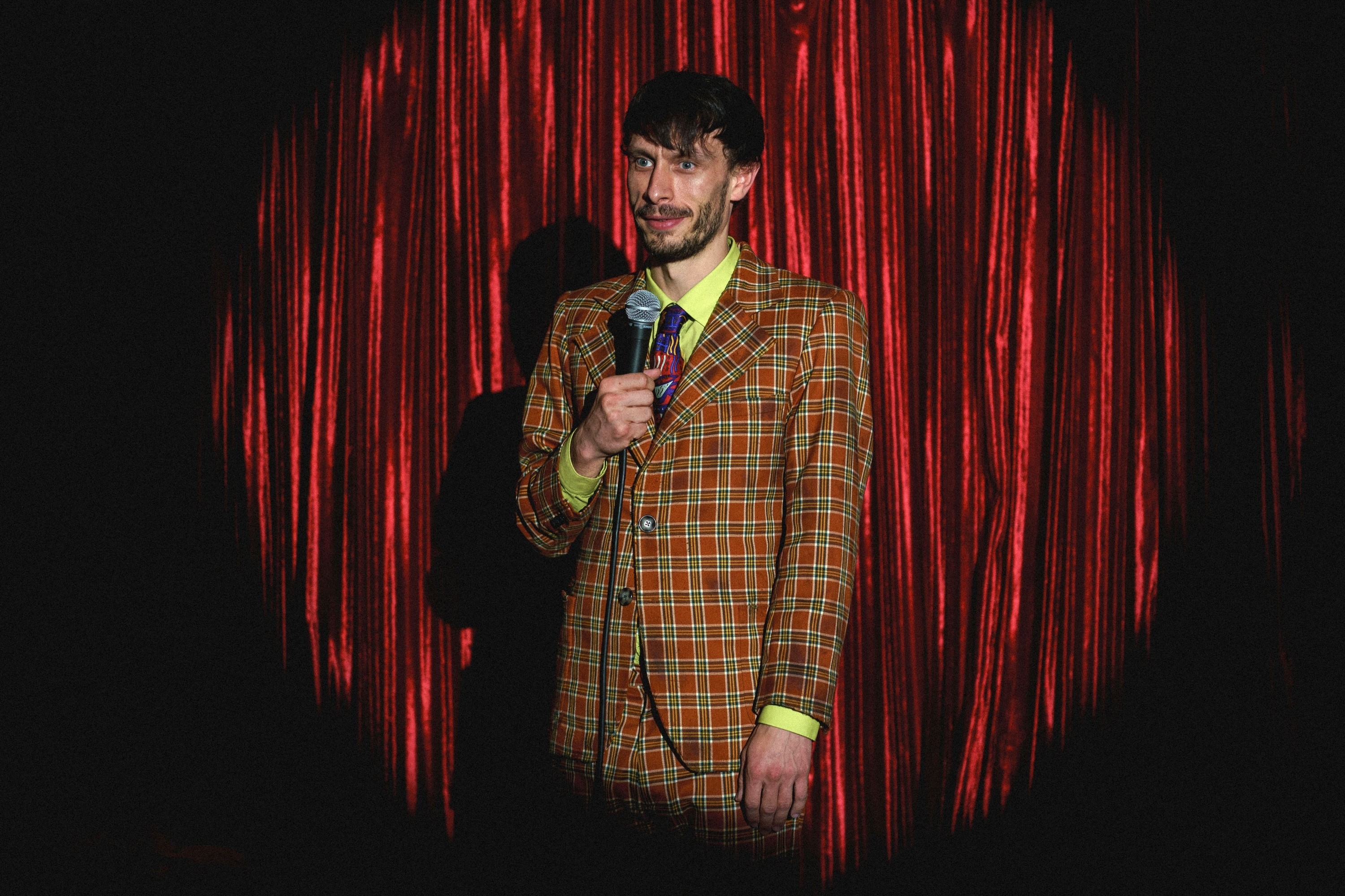 Richard Gadd in a yellow-and-red plaid suit holding a microphone with a spotlight on him and red curtain in the background