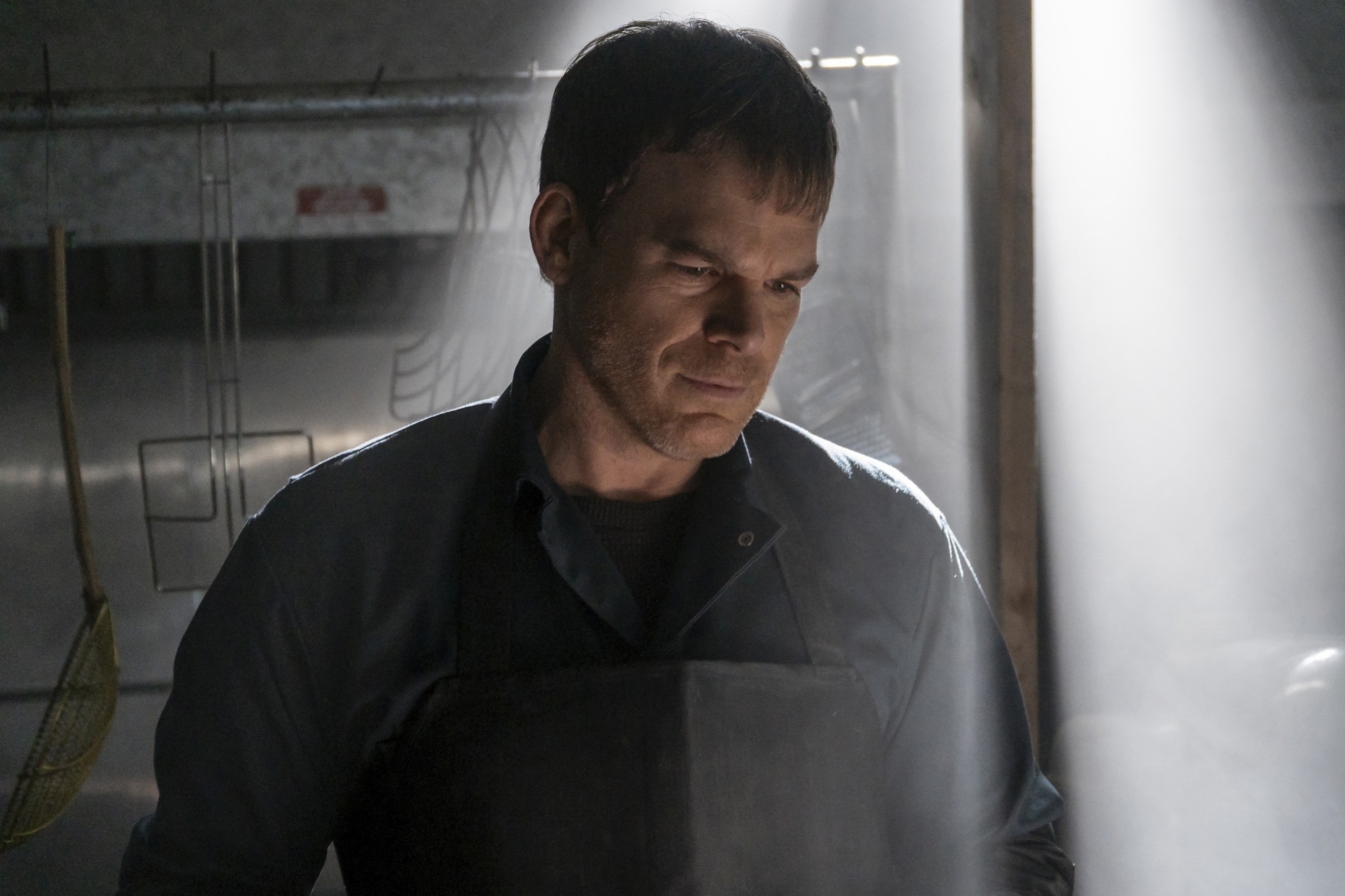 Michael C. Hall as Dexter Morgan looks down thoughtfully, wearing a black apron over a blue shirt, bathed in dramatic lighting