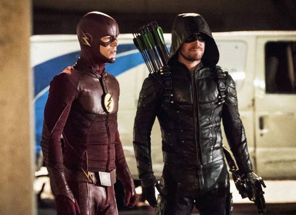 Grant Gustin (as The Flash), Stephen Amell (as Green Arrow) standing in front of a white vehicle with a blue pattern on it during the evening hours