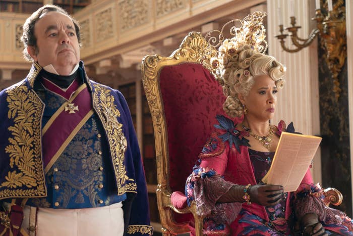 Paul Ritter stands in a regal blue uniform next to a seated Golda Rosheuvel dressed in an elaborate royal gown with an intricate wig