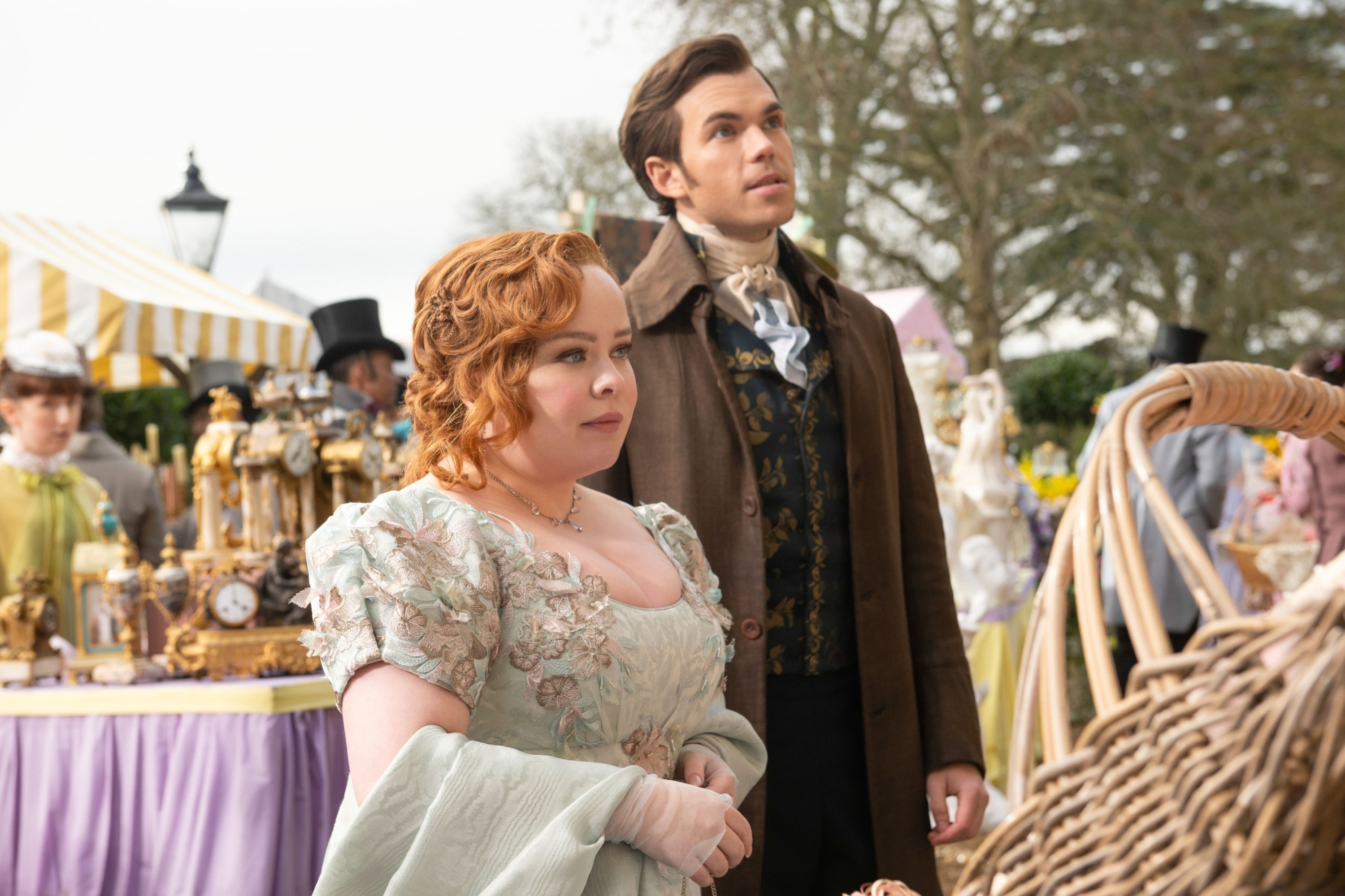 Nicola Coughlan and Luke Newton in period costumes at a market scene from "Bridgerton." Coughlan wears a light dress with floral details; Newton in a dark coat