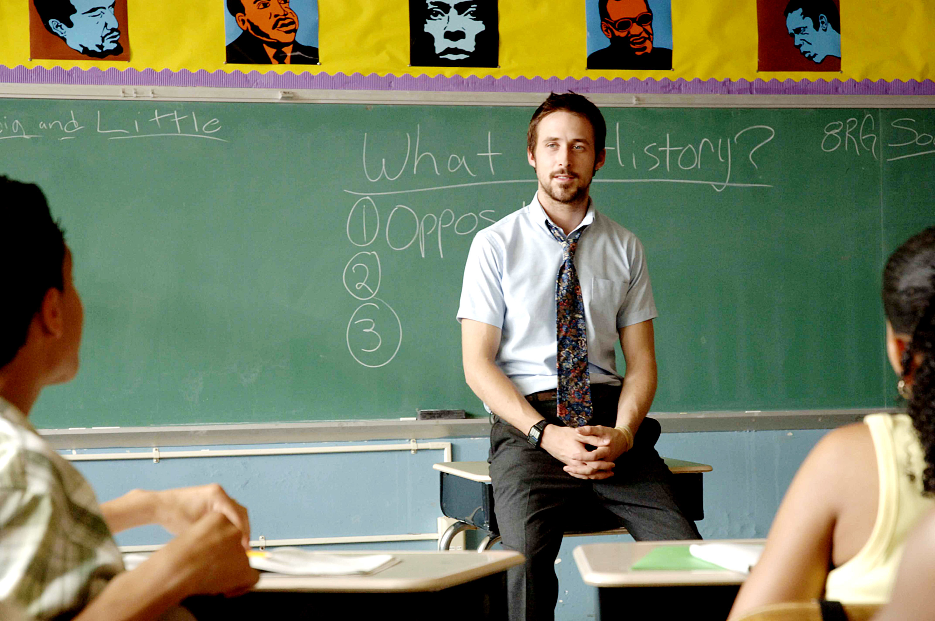 Ryan Gosling, seated on a desk, talks to students in a classroom with a green chalkboard and posters of famous leaders on the wall