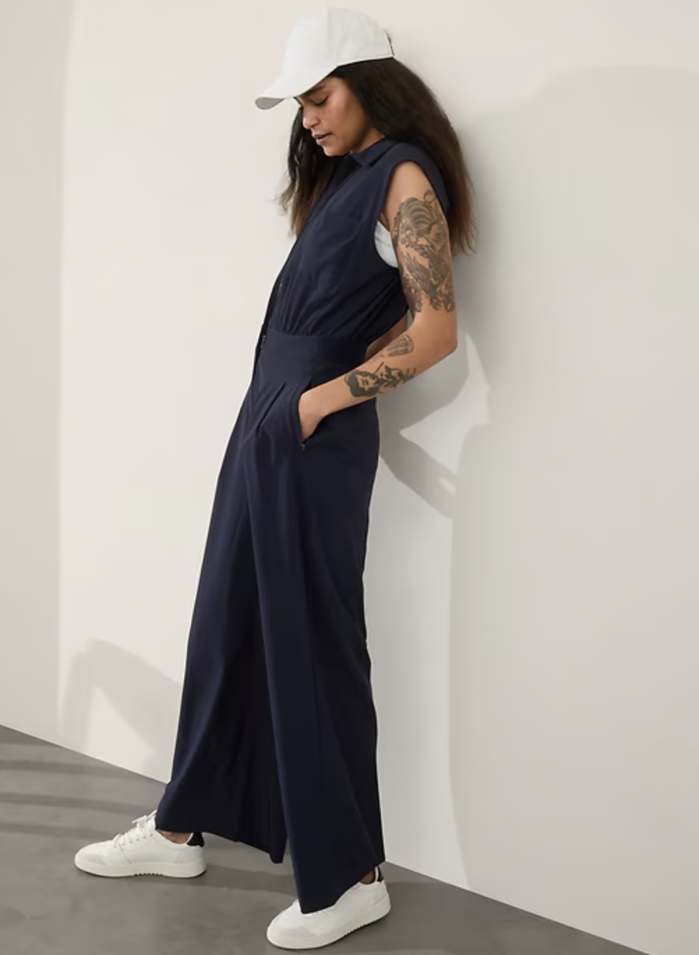 Model leaning against a wall, wearing a sleeveless jumpsuit, white cap, and white sneakers, showing tattoos on their arm