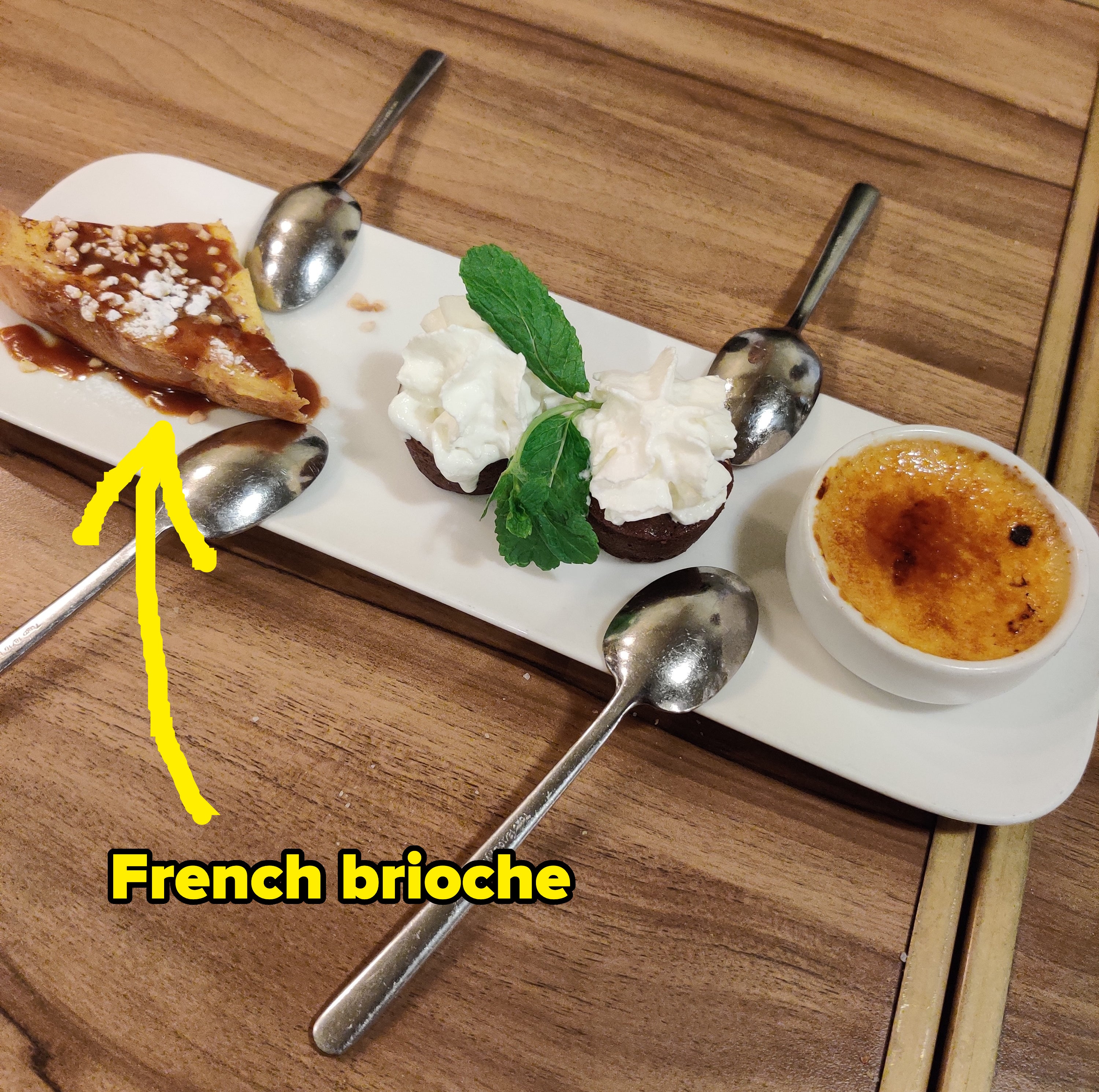 A dessert platter featuring caramel-topped pastry, whipped cream with mint leaves, and a small creme brulee with four spoons on a wooden table
