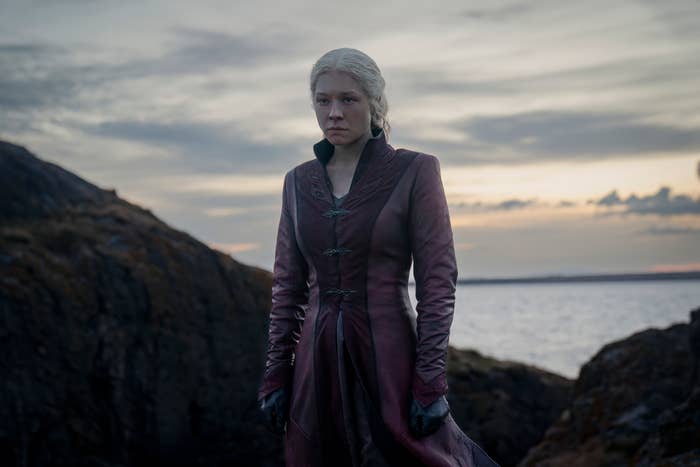 Emma D'Arcy as Rhaenyra Targaryen wears a dark medieval-style outfit, standing on a rocky seaside landscape, from the TV show "House of the Dragon."