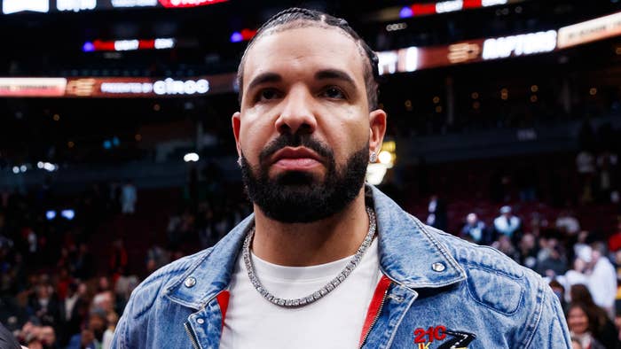 Drake stands in a crowd at an event, wearing a jean jacket over a white shirt, with a chain necklace