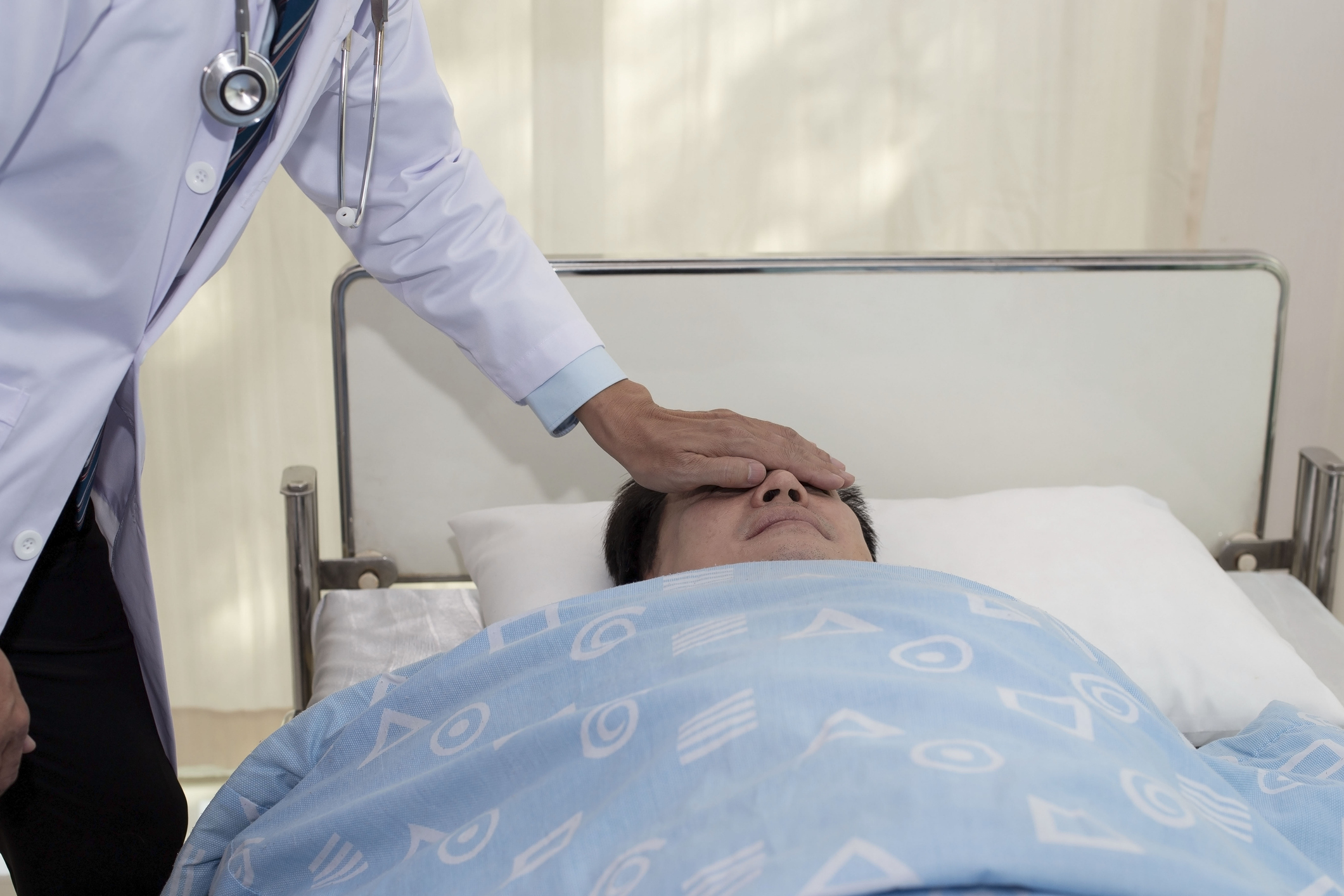A doctor places a hand on the forehead of a patient lying in a hospital bed, covered with a blanket