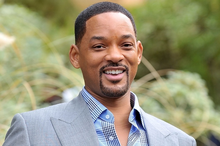 Will Smith smiling in an outdoor setting, wearing a light gray suit with a blue checkered shirt