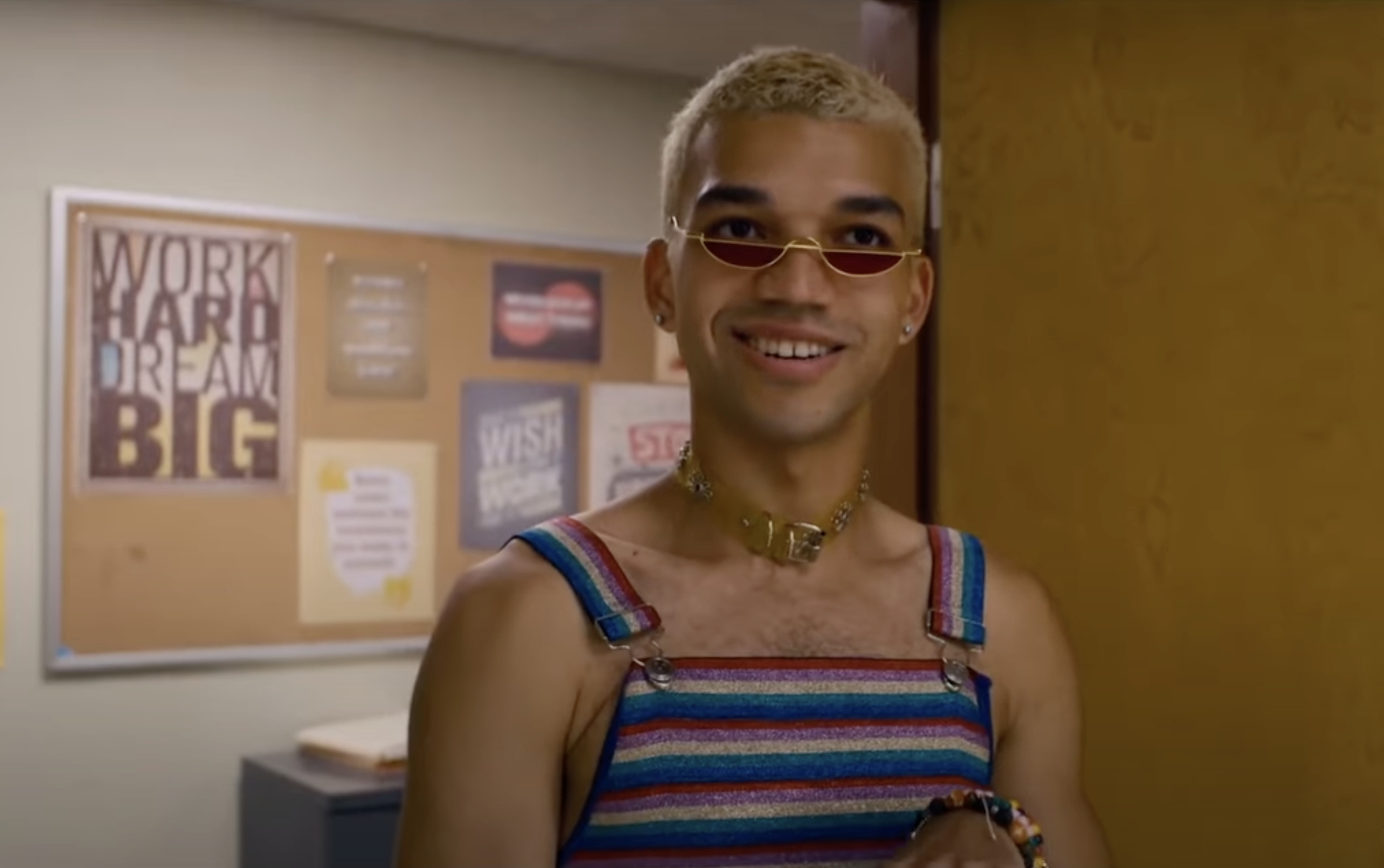 Justice Smith wearing a striped sleeveless top, small sunglasses, and gold jewelry, smiles in front of a bulletin board with motivational posters
