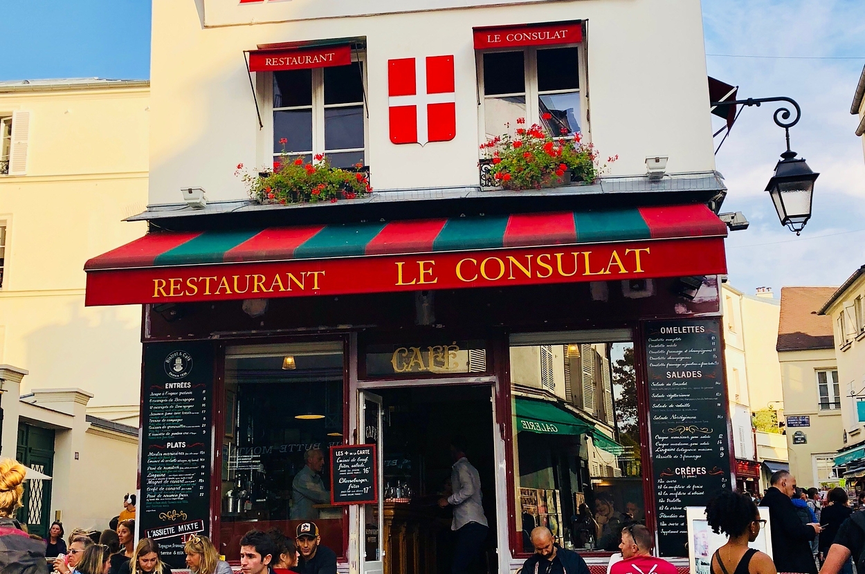 Exterior of Le Consulat restaurant in Paris with patrons sitting at outdoor tables, captured in a lively street scene