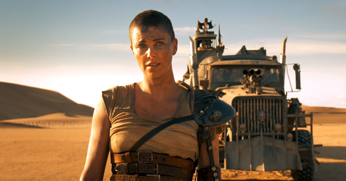 Charlize Theron as Furiosa stands in front of a post-apocalyptic vehicle in a desert setting, wearing a rugged, battle-worn outfit in "Mad Max: Fury Road"