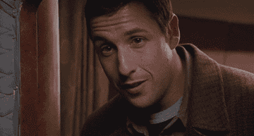 Adam Sandler winks and smiles while standing indoors, wearing a casual jacket over a T-shirt