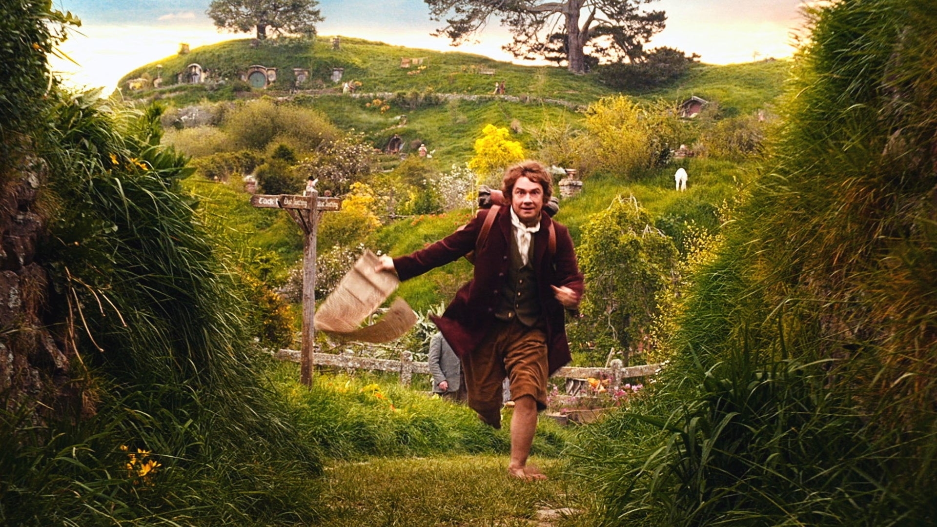 Bilbo Baggins, played by Martin Freeman, runs energetically in a lush green Hobbiton landscape, carrying a burlap sack
